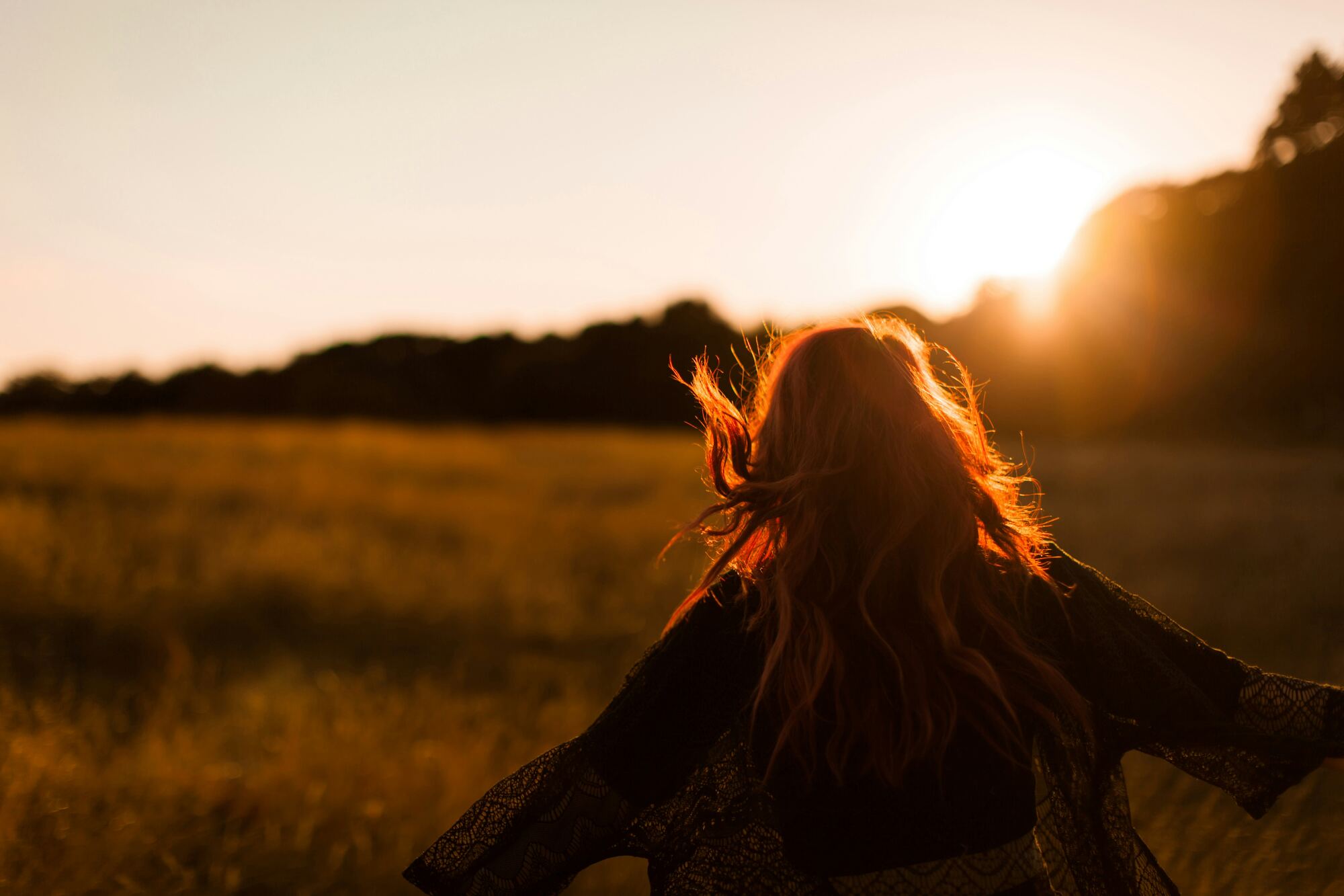 A woman with long hair walks through an open field at sunset, arms outstretched in the golden light